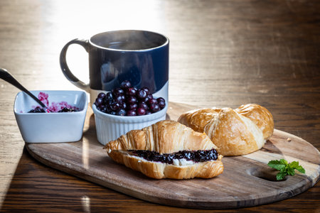 A plate of croissants with huckleberry jam and a cup of coffee sit on a wooden table. The croissants are arranged on a wooden tray, and the coffee is in a blue mug. Concept of comfort and relaxationの写真素材