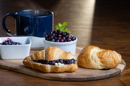 A plate of croissants with huckleberry jam and a cup of coffee sit on a wooden table. The croissants are arranged on a wooden tray, and the coffee is in a blue mug. Concept of comfort and relaxationの写真素材