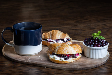 A plate of croissants with huckleberry jam and a cup of coffee sit on a wooden table. The croissants...