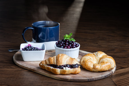 A plate of croissants with huckleberry jam and a cup of coffee sit on a wooden table. The croissants are arranged on a wooden tray, and the coffee is in a blue mug.の写真素材