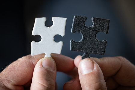 A hand holding two puzzle pieces, one white and one black both pieces have the same shape. The...