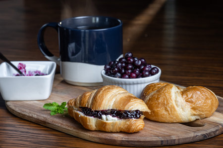 A plate of croissants with huckleberry jam and a cup of coffee sit on a wooden table. The croissants...