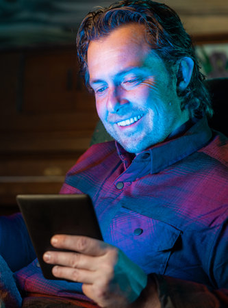 A man is sitting in a chair and smiling while looking at a tablet reading a digital book. He is...