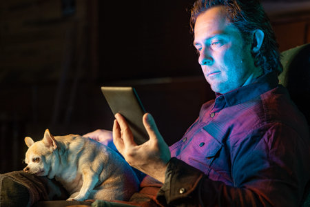 A man is sitting in a chair with a dog on his lap. He is holding a tablet in his hand reading a book.
