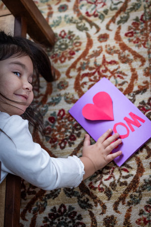 A little girl is in the furniture looking at a card that says "Mom". She is smiling and seems to be happyの写真素材