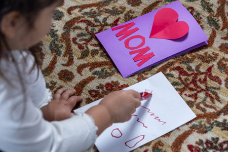 A little girl is drawing a card for her mom. She is using a red marker to write the words Mom on the...