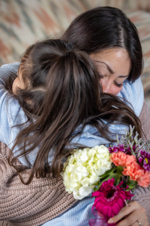 A young mother is holding a bouquet of flowers from her little girl as a Mother's Day present. The woman is smiling and the girl is looking at her. The flowers are pink and whiteの写真素材