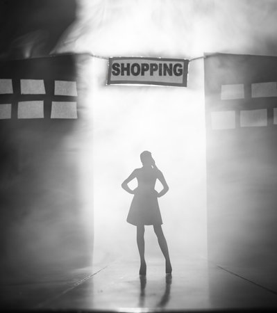 Creative conceptual image of a female silhouette Standing with her Arms on her Hips in Front of a city with a Banner that Reads SHOPPING. The scene features dramatic lighting, theatrical smoke, and cardboard cutouts representing buildings and storefronts.の写真素材