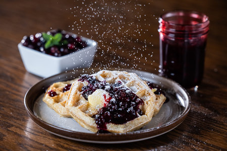 Close-up of golden waffles topped with butter, fresh huckleberries, and berry jam as powdered sugar is sprinkled on top. Rustic breakfast or dessert food photography with a gourmet, homemade style.の写真素材