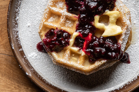 Close-up of golden waffles topped with butter, fresh huckleberries, and berry jam as powdered sugar is sprinkled on top. Rustic breakfast or dessert food photography with a gourmet, homemade style.の写真素材