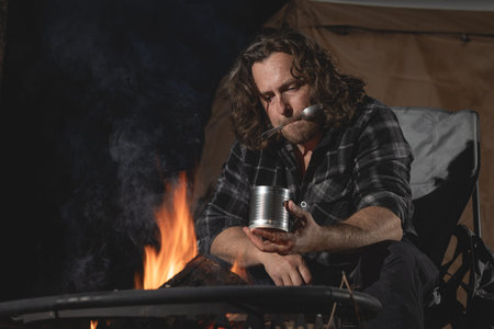 A man camping sitting by the fire eating canned food.