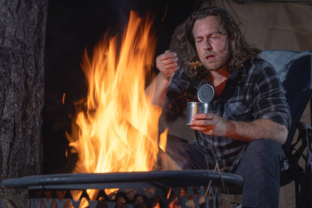 A man camping sitting by the fire eating canned food.