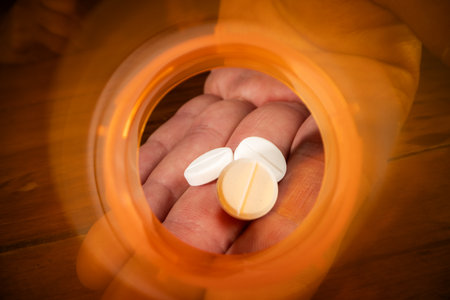 Dramatic macro view looking from the inside of an empty, bright orange prescription pill bottle. A human hand is visible reaching a small dose of tablets.の写真素材