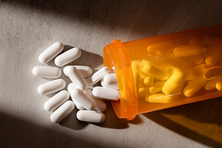 A photograph of capsules and pills spilling from an orange bottle onto a concrete surface,...