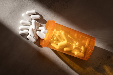 A dramatic, low-key photograph of capsules and pills spilling from an orange bottle onto a concrete...