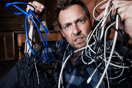 A stressed adult male struggles with a chaotic tangle of various electronic cables, USB cords, and...