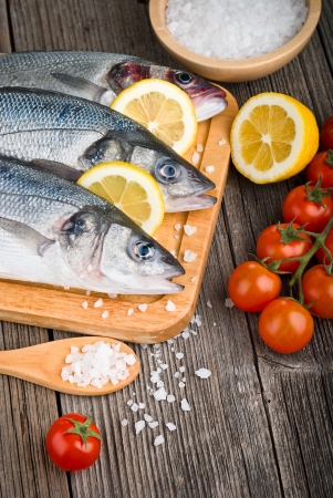 Fresh sea bass on chopping board with lemon and vegetables on wooden tableの写真素材