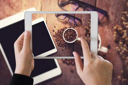 hand girl holding tablet to take photo of cup of coffee on wood tableの写真素材