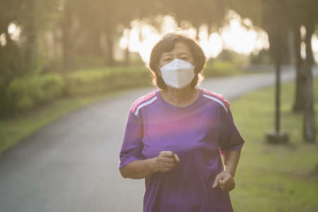 Athletic asian senior woman 60s wearing surgical mask and jogging. Beautiful senior asian woman running at the park on a sunny day.の写真素材