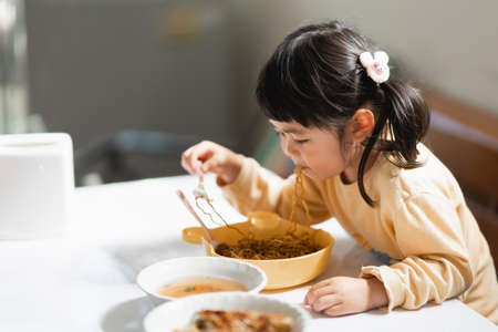 Asian baby eating delicious noodle in kitchen on dining table. Happy asian baby girl practice eating by her self on dining table. Baby lifestyle conceptの写真素材