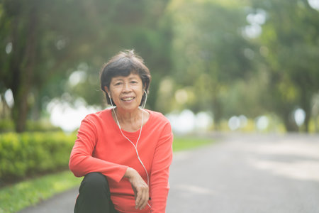 senior asian woman runner smiling use mobile smart phone app tracking progress listening to music with earphones for fitness motivation. Asian woman sitting prepare run at parkの写真素材