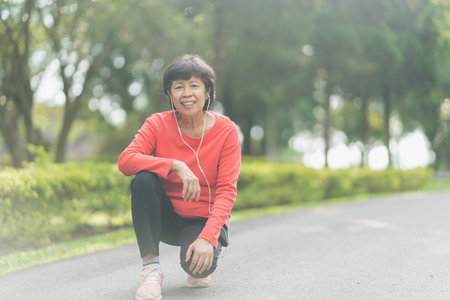 senior asian woman runner smiling use mobile smart phone app tracking progress listening to music with earphones for fitness motivation. Asian woman sitting prepare run at parkの写真素材