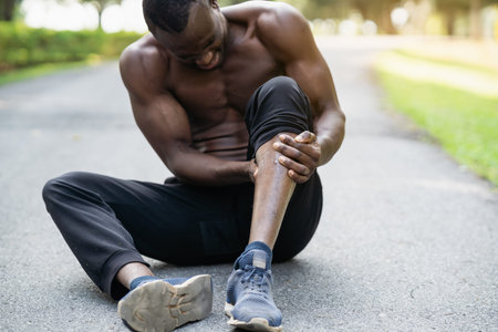 African runner man sitting on jogging track and feeling pain in his leg, copy space. Sports injury...