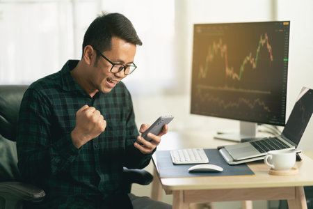 Asian man trader drinking coffee and sitting at home office in front of monitors with cryptocurrency graph holding smartphone browsing application monitoring cryptocurrency priceの写真素材