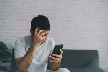 Depression concept. Asian man sad and checking mobile phone sitting on the sofa in the living room at home, Asian man serious and worry while using mobile phone.の写真素材