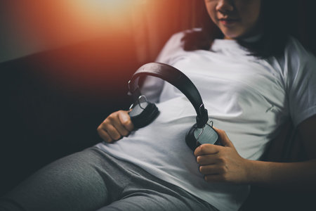 Pregnant woman putting headphones on her belly while lying on a bed at home. Family mother mom...