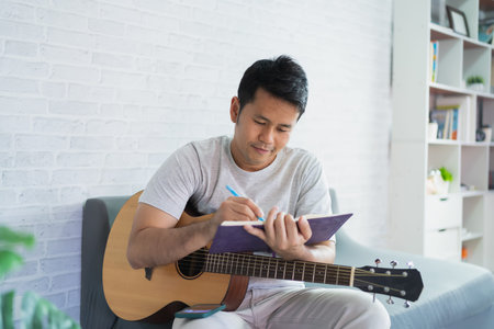 Asian man playing guitar while sitting on sofa in light living room. Asian man writing song while playing guitar at home. Compose song music concept.の写真素材