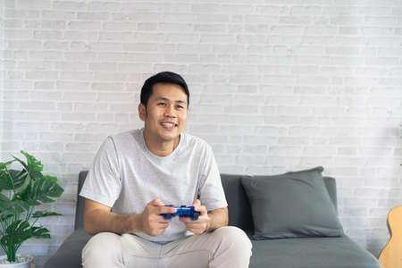 Smiling asian man wearing casual clothes sitting on a couch at the living room. Asian man using mobile phone chatting in living room.の写真素材