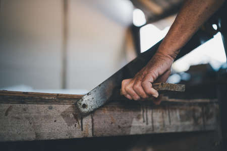 Senior old carpenter using saw working on wood craft at workshop to produce construction material or wooden furniture.の写真素材