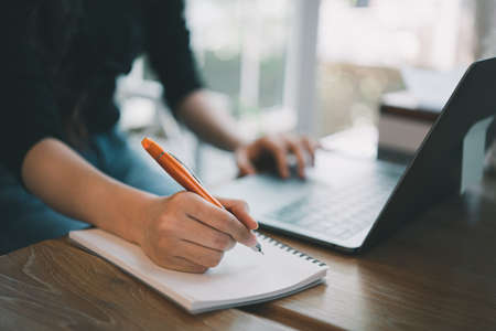 closeup hands of business woman working on laptop and using pen making notices in her notebook on wooden table with a cup of coffee. Business conceptの写真素材