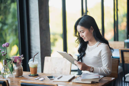 Asian woman typing text message on tablet in a cafe. Young Asian woman sitting at a table with a coffee using tablet trading stock market or crypto currencyの写真素材
