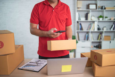 Delivery man worker smiling using mobile scan barcode for boxes for sending or conveying parcels by mail. Wearing red uniform work at home.の写真素材