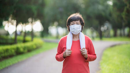 Athletic asian senior woman 60s wearing surgical mask and jogging. Beautiful senior asian woman running at the park on a sunny day.の写真素材