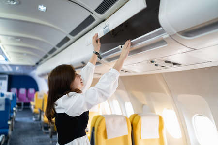 Asian young woman open the top shelf to get luggage on airplane.Cabin crew girl lift luggage bag in airplane . Airline transportation and tourism concept.の写真素材