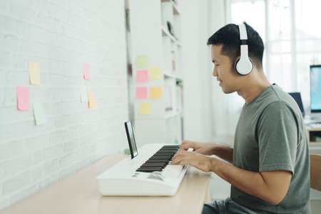Asian man wearing white headphones play piano and sing a song and learning online with mobile phone and compose writing song or record sound with a microphone and use computer recording music programの写真素材