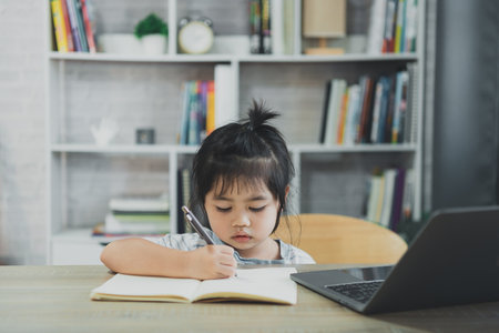 Asian baby girl wearing a blue striped shirt use laptop and notebook to study online on wood table desk in living room at home. Education learning online from home concept.の写真素材