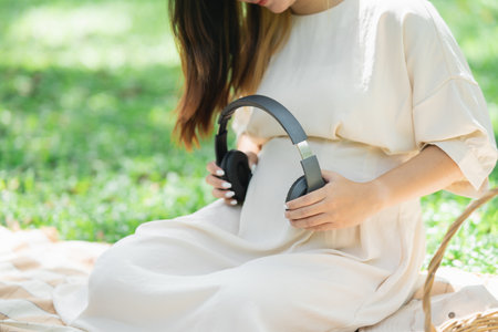 Pregnant happy asian woman putting headphones on her belly to listen music while sitting on picnic blanket on lawn at the garden park under the tree. Family mother mom pregnant healthy concept.の写真素材