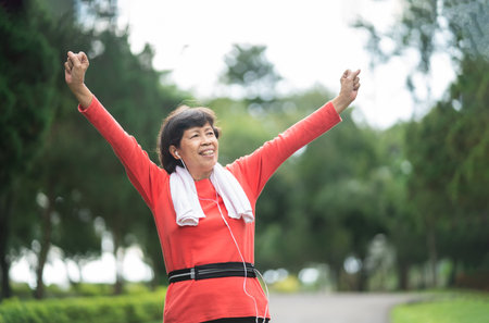 Athletic asian senior woman 60s smiling, hands up and happy cheerful with success and jogging. Senior asian woman running at park. Female runner listening to music with earphones while jogging.の写真素材