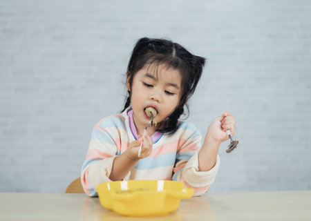 Asian baby girl happy using cutlery spoon and fork eating delicious noodle and meatball in kitchen on dining table. Happy asian baby girl practice eating by her self on dining table. Baby food conceptの写真素材