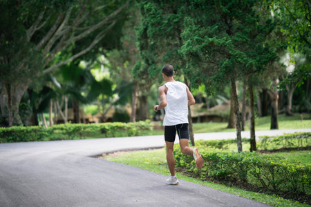 Back side view. Asian man sportsman fit body wearing smart watch while running or jogging in the garden have more tree and clean air, exercising at park. Sport healthy running concept.の写真素材