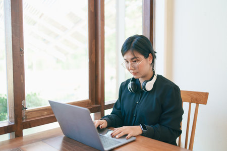 Business freelance entrepreneur woman wearing eyeglass and white headphone working typing keyboard on laptop and on wooden table at the cafe coffee shop. Business technology concept.の写真素材