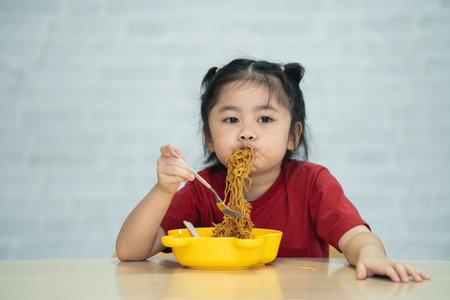 Asian baby girl enjoy happy using cutlery spoon and fork eating delicious noodle in kitchen on dining table. Happy asian baby girl practice eating by her self on dining table. Baby food conceptの写真素材