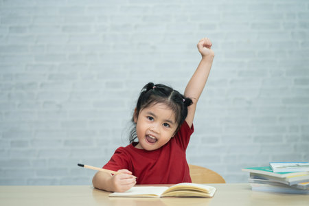 Asian baby girl wearing a red shirt  write notes in notebook and reading book to study online and raise your hand on wood table desk in living room at home. Education learning online from home conceptの写真素材