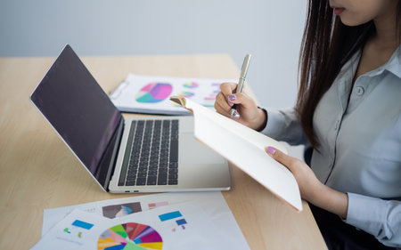 Asian young woman in smart casual wear writing on notebook and working on laptop while sitting in creative office or cafe. young girl working with laptop on the wood table in the cafeの写真素材