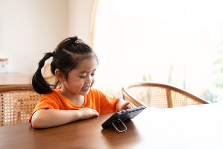 Asian child girl looking using and touch mobile phone screen. Baby smiling funny time to use mobile phone. Too much screen time. Cute girl watching videos while tv, Internet addiction concept.の写真素材