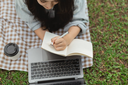 Top view. Asian woman lay on picnic blanket and lawn at park garden working on laptop and writing on notebook. Asian female using laptop while sitting under a tree at park. Work from anywhere concept.の写真素材